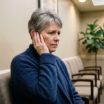 Femme d'une cinquantaine d'années dans une salle d'attente médicale tenant son oreille avec une expression préoccupée