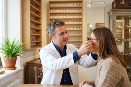 Opticien indépendant ajustant des lunettes sur une cliente dans une boutique artisanale aux étagères en bois garnies de montures